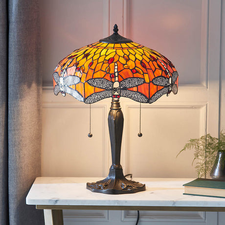 Tiffany Alderworth medium table lamp in flame, amber leaded glass shade with dragonfly motif on a turned brass-finish base, shown on a marble console.