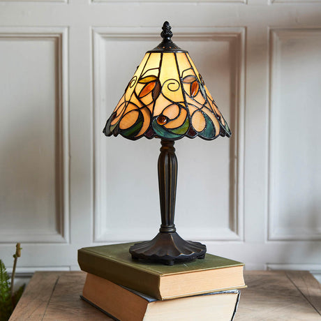 Larncombe Mini Tiffany table lamp with multicoloured leaded glass shade and dark metal base, displayed on stacked books against a panelled wall.