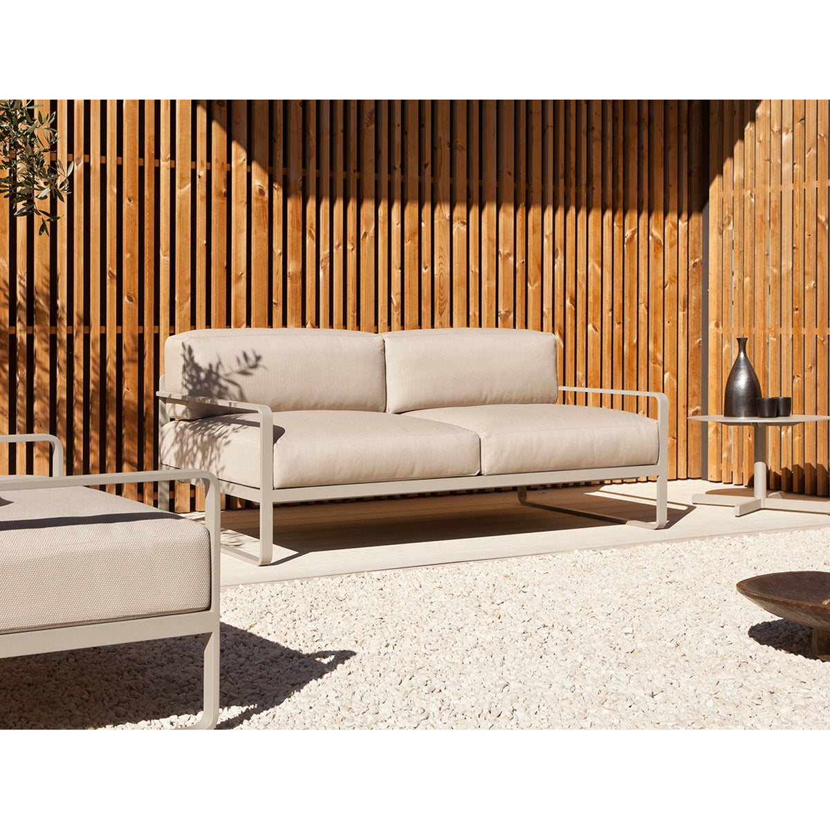 Sit Aluminium 2 Seater Garden Sofa with beige cushions and white powder-coated sled-leg frame, shown on a gravel patio against a wooden slat wall.