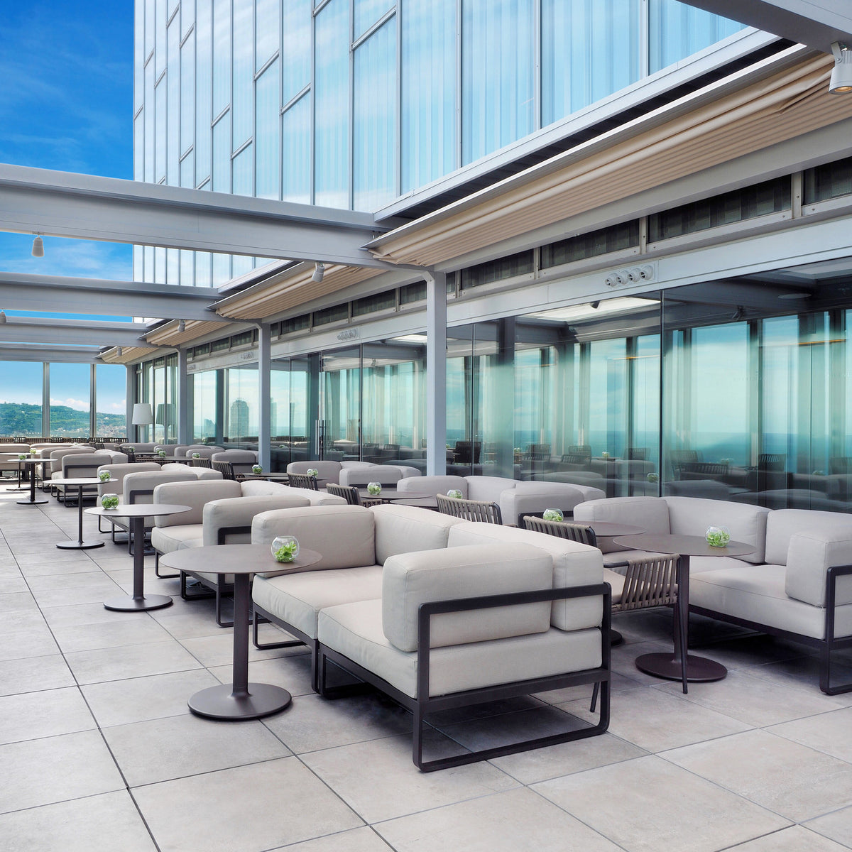 Sit Aluminium 2 Seater Garden Sofa with cream cushions and dark frame, shown on a rooftop terrace with round side tables and glass balustrade.