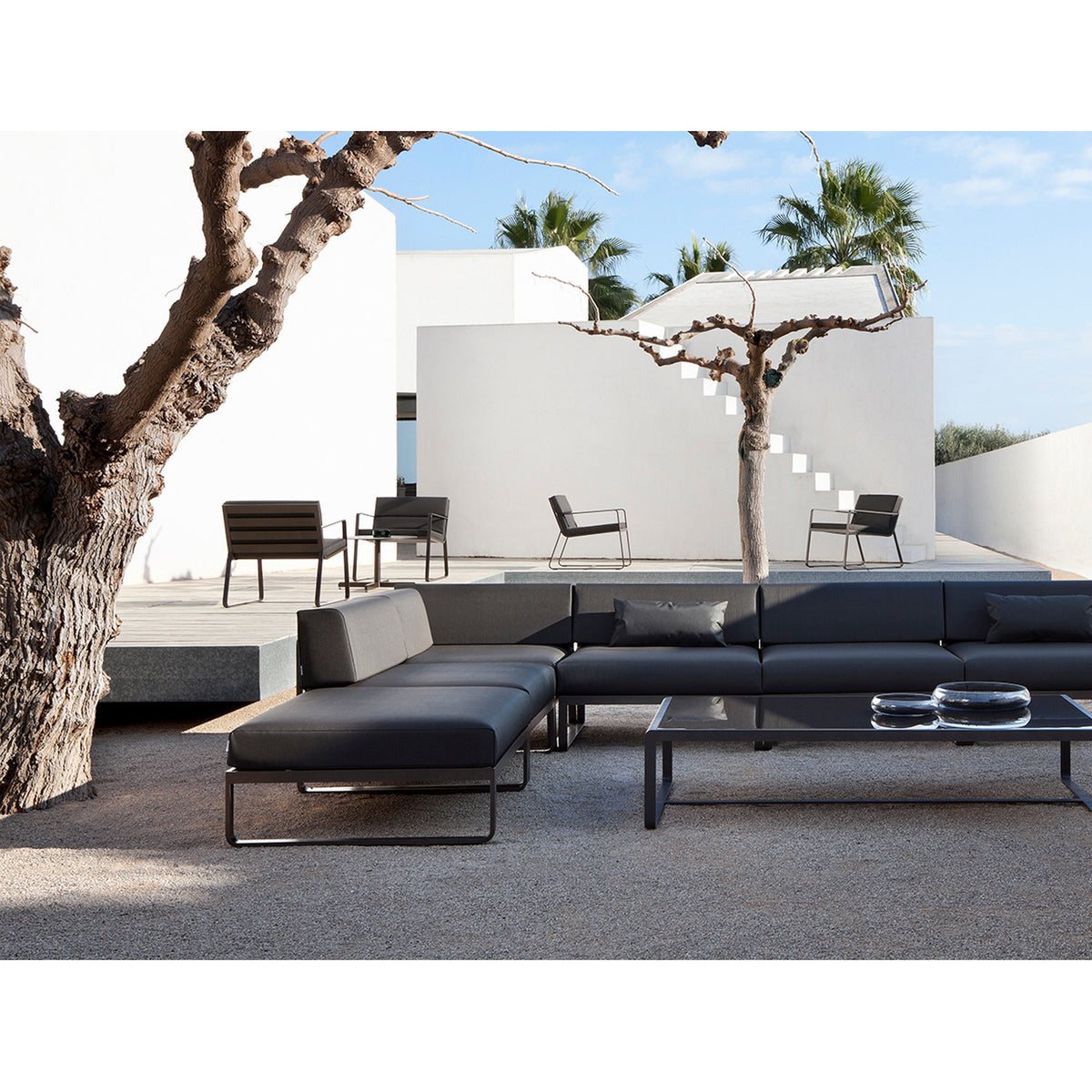 Sit aluminium garden corner sofa in black with grey cushions, shown as part of a modular outdoor seating arrangement on a stone patio beside a tree and white villa.