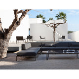 Sit aluminium garden corner sofa in black with grey cushions, shown as part of a modular outdoor seating arrangement on a stone patio beside a tree and white villa.