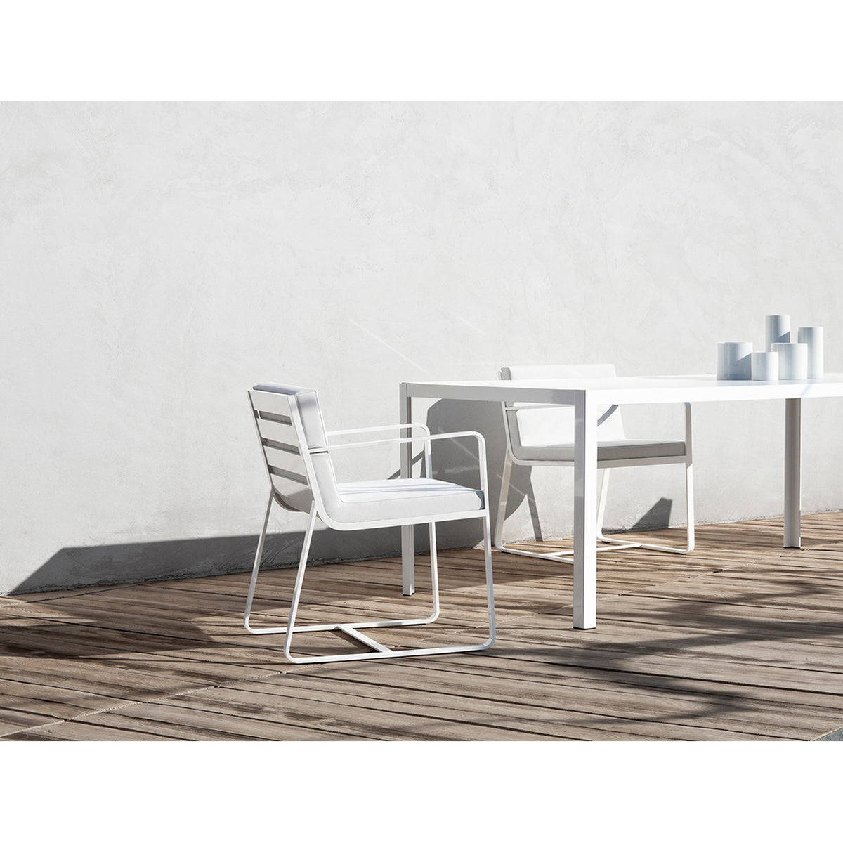 White aluminium Sit garden dining armchair with slatted back, flat arms and sled legs, shown with cushions on a patio beside a matching table.