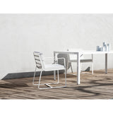 White aluminium Sit garden dining armchair with slatted back, flat arms and sled legs, shown with cushions on a patio beside a matching table.