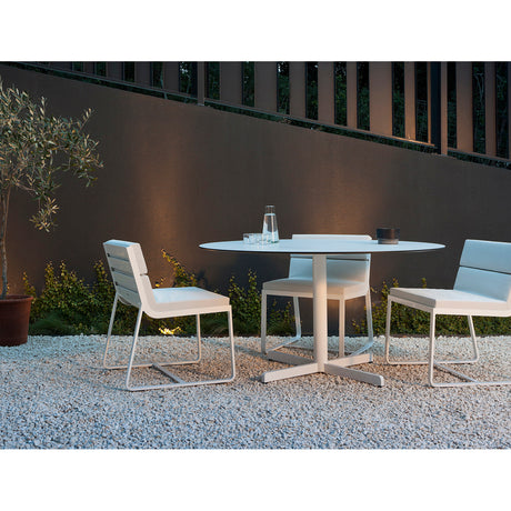 Sit aluminium garden dining chairs with white cushions and sled-leg frames, shown around a round white outdoor table on gravel against a dark wall.