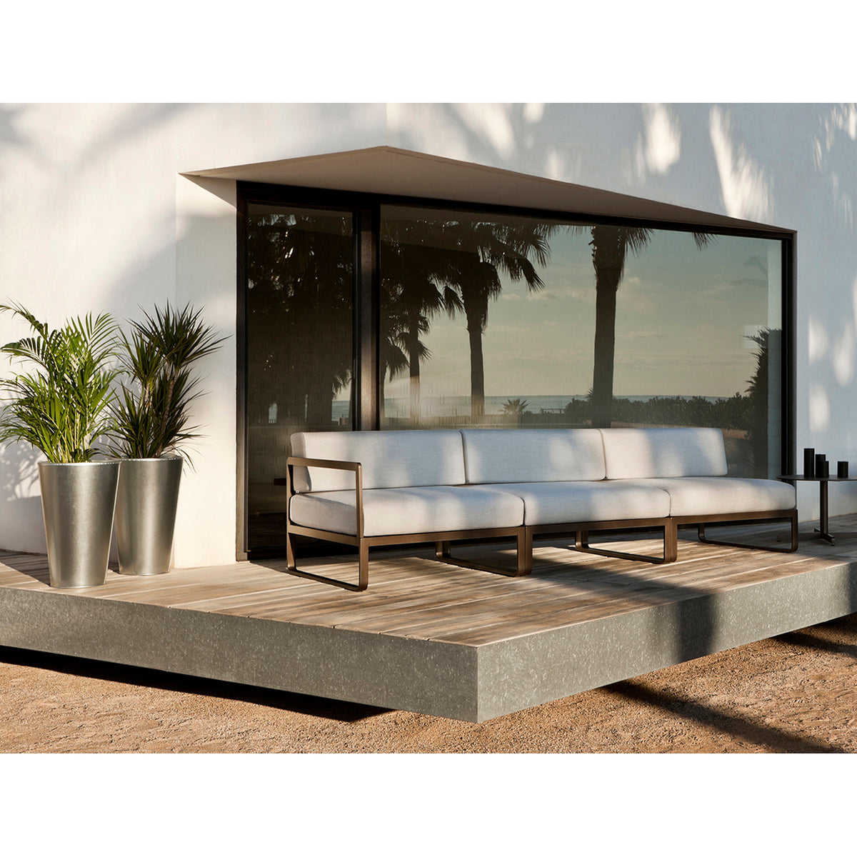 Sit Aluminium Modular Garden Sofa with right arm module, white cushions and bronze-finished aluminium frame, shown on a wooden deck beside a glass-fronted outdoor room.