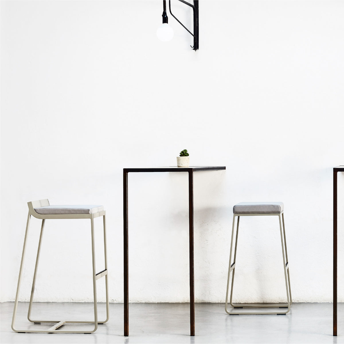 Sit aluminium outdoor bar stools with low backrest and grey cushioned seats, sled-leg frames with footrest bars, shown beside a tall table against a white interior wall.