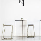 Sit aluminium outdoor bar stools with low backrest and grey cushioned seats, sled-leg frames with footrest bars, shown beside a tall table against a white interior wall.
