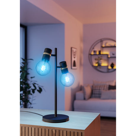 Two Smart Colour Changing LED Globe bulbs glowing blue with visible spiral filaments, fitted to a black two-head table lamp in a living room setting.