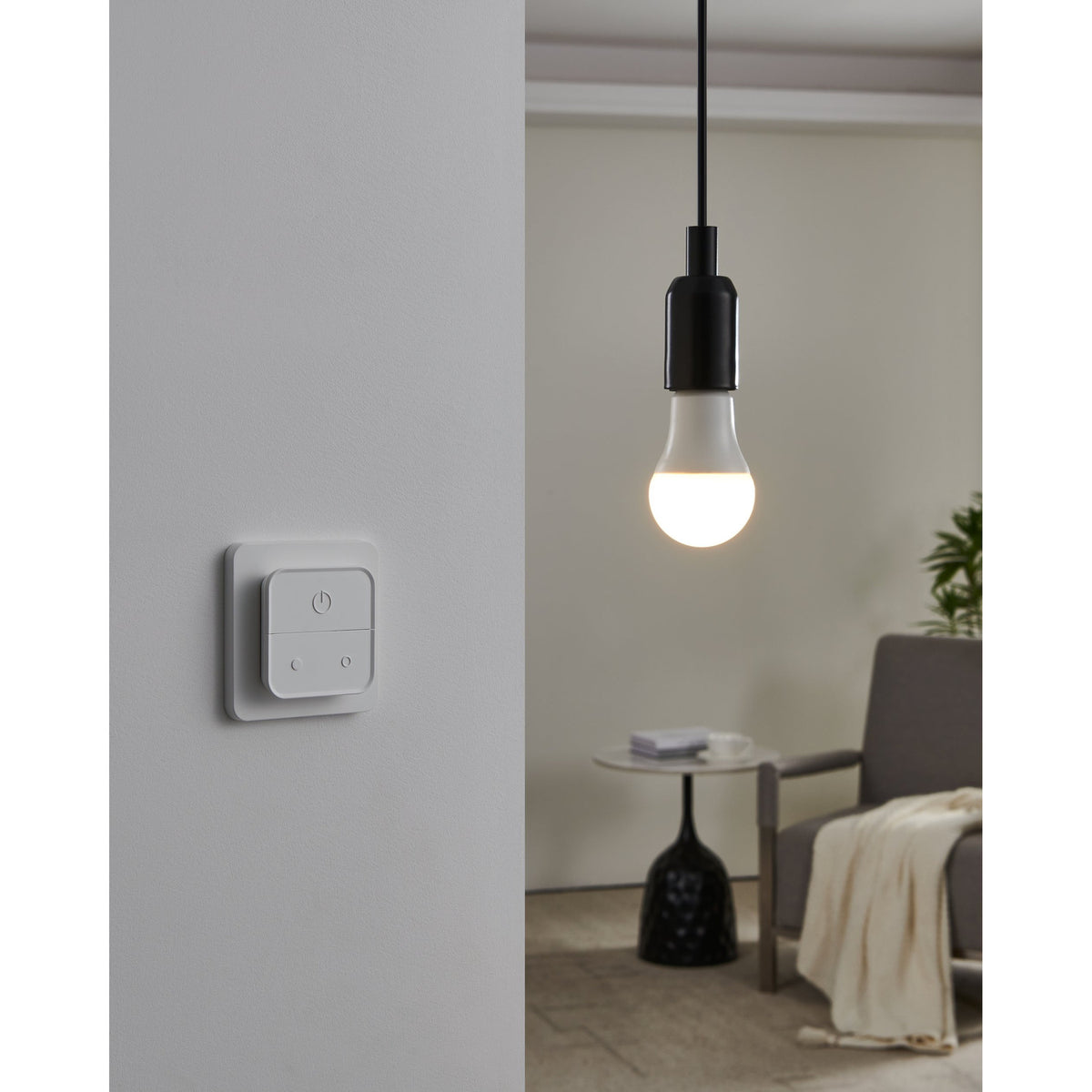 Smart Colour Changing LED GLS Bulb E27, warm-lit opal globe in a black pendant fitting above a living room armchair and side table, controlled by a white wall-mounted smart switch.