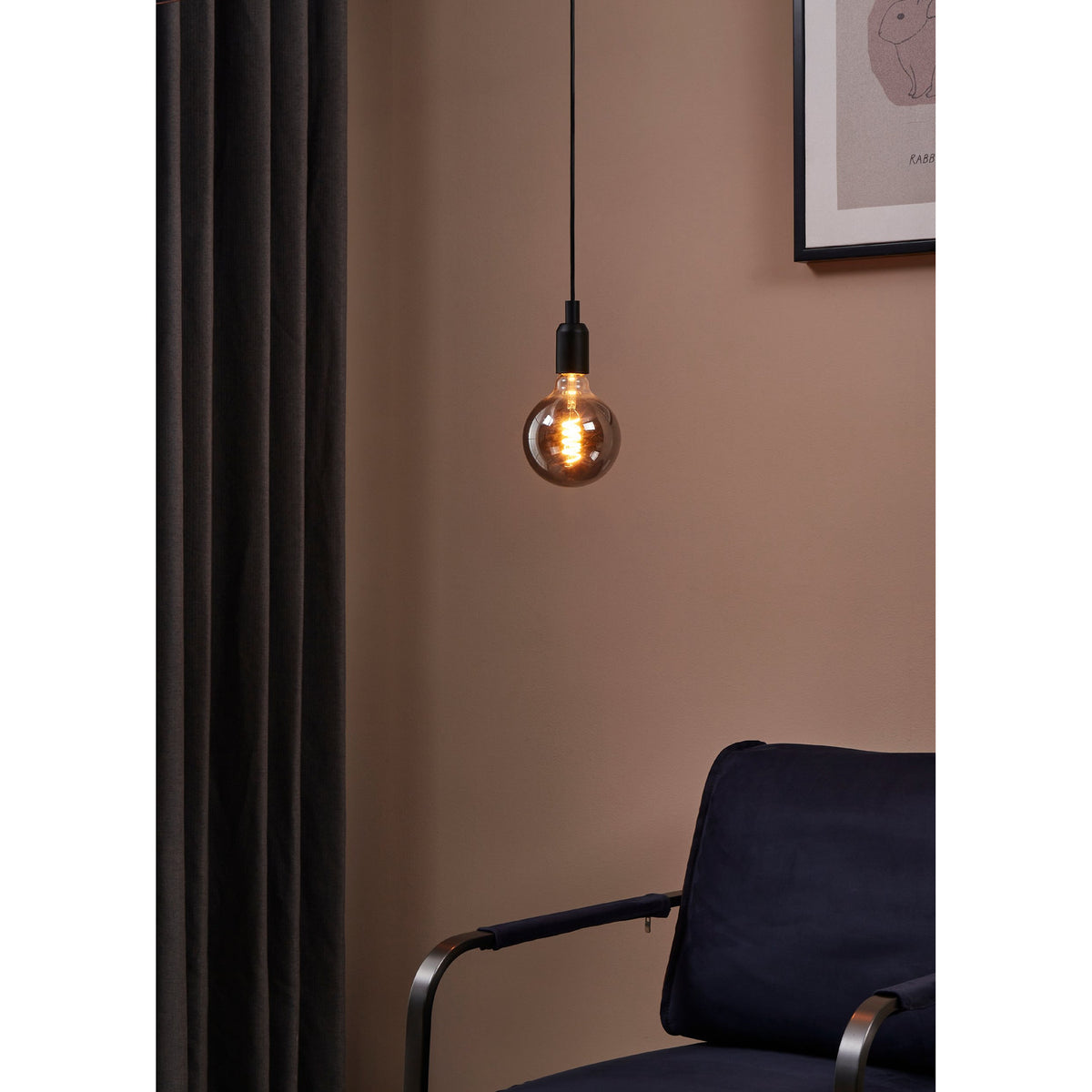 Smoked LED globe pendant bulb with E27 screw cap and spiral filament, glowing warm white on a black cord fitting against a pink wall in a living room setting.