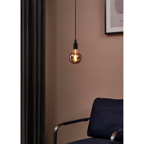 Smoked LED globe pendant bulb with E27 screw cap and spiral filament, glowing warm white on a black cord fitting against a pink wall in a living room setting.