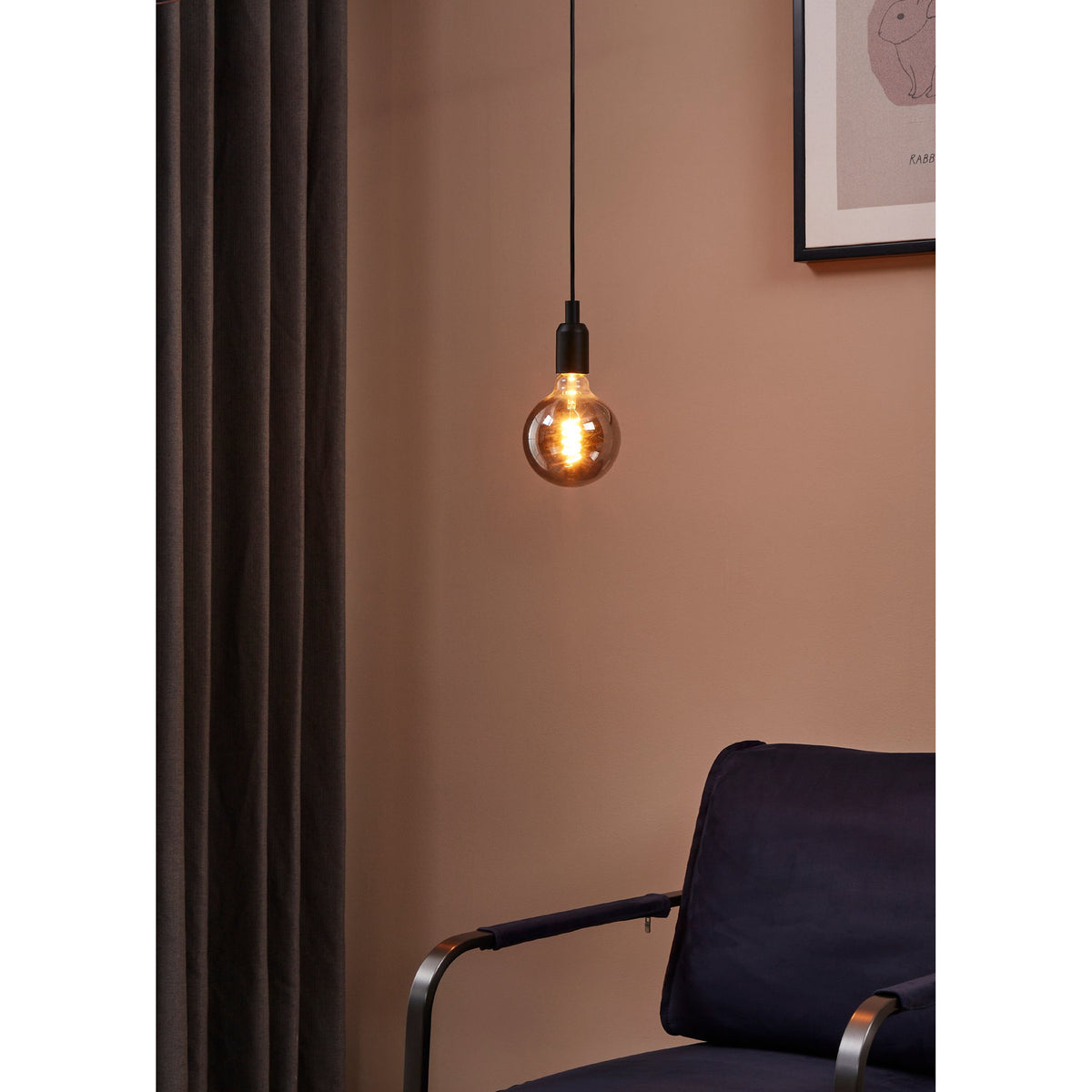 Smoked LED globe bulb with visible spiral filament, E27 fitting, hung from a black pendant cord against a pink wall in a living room with a dark curtain and navy armchair.