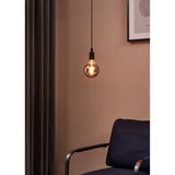 Smoked LED globe bulb with visible spiral filament, E27 fitting, hung from a black pendant cord against a pink wall in a living room with a dark curtain and navy armchair.