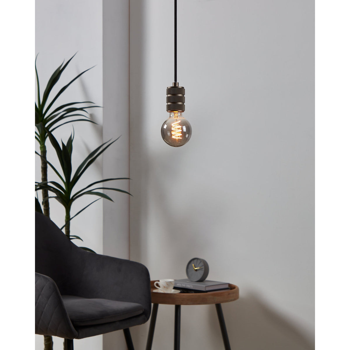 Smoked glass LED globe bulb with visible spiral filament on a pendant fitting with bronze finish, shown lit in a living room beside a dark chair and side table.
