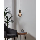 Smoked glass LED globe bulb with visible spiral filament on a pendant fitting with bronze finish, shown lit in a living room beside a dark chair and side table.