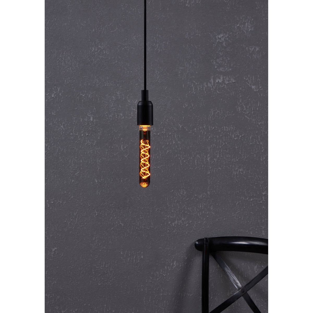 Smoked LED tubular bulb E27 with glowing amber spiral filament, hanging from a black pendant fitting against a dark textured wall.