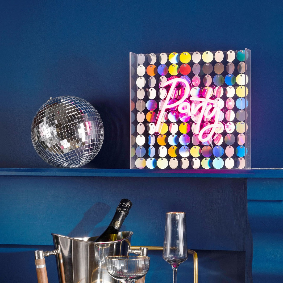 Party Neon Sign in pink script on a square sequin panel, displayed on a blue shelf beside a disco ball in a living room setting.