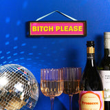 Black-framed rectangular wooden hanging plaque with gold inner edge and hot pink panel reading "BITCH PLEASE" in bold yellow capitals, shown on a blue wall above a disco ball and drinks.