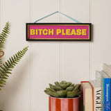 Rectangular wooden hanging plaque with black frame and gold inner edge, reading "BITCH PLEASE" in bold yellow capitals on a hot pink panel, shown hung on a wall above a shelf of books and a plant.