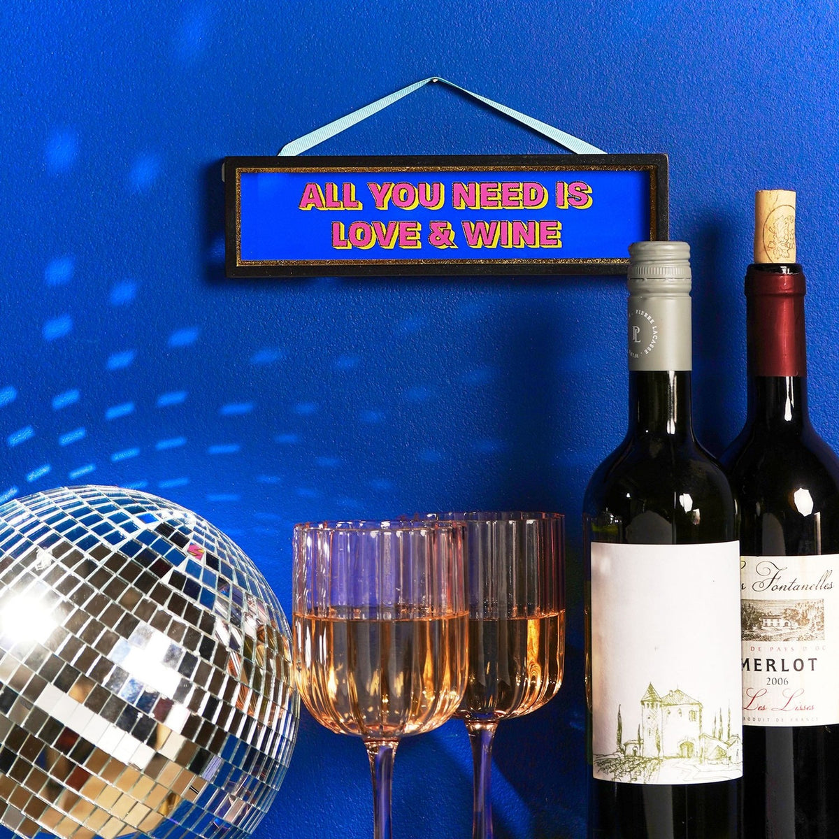 Black-framed wooden hanging plaque reading "All You Need Is Love & Wine" in pink and yellow on blue, shown on a wall beside wine bottles, glasses, and a disco ball.