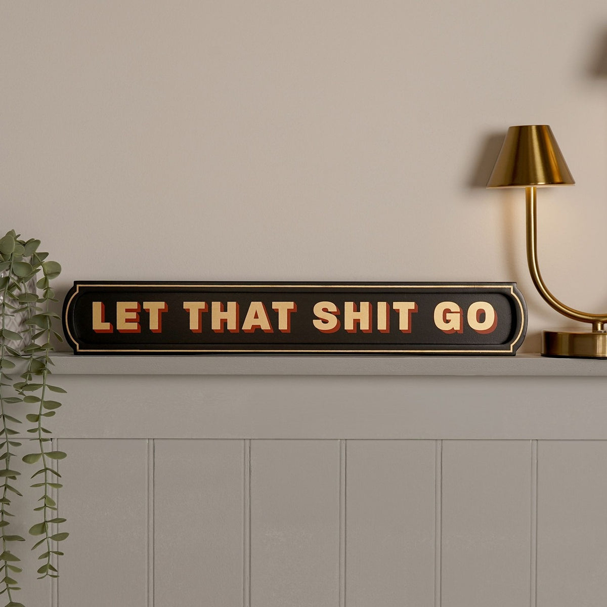 Black wooden wall plaque reading "LET THAT SHIT GO" in bold cream letters with red shadow, shaped like a vintage sign, displayed on a shelf beside a brass lamp.