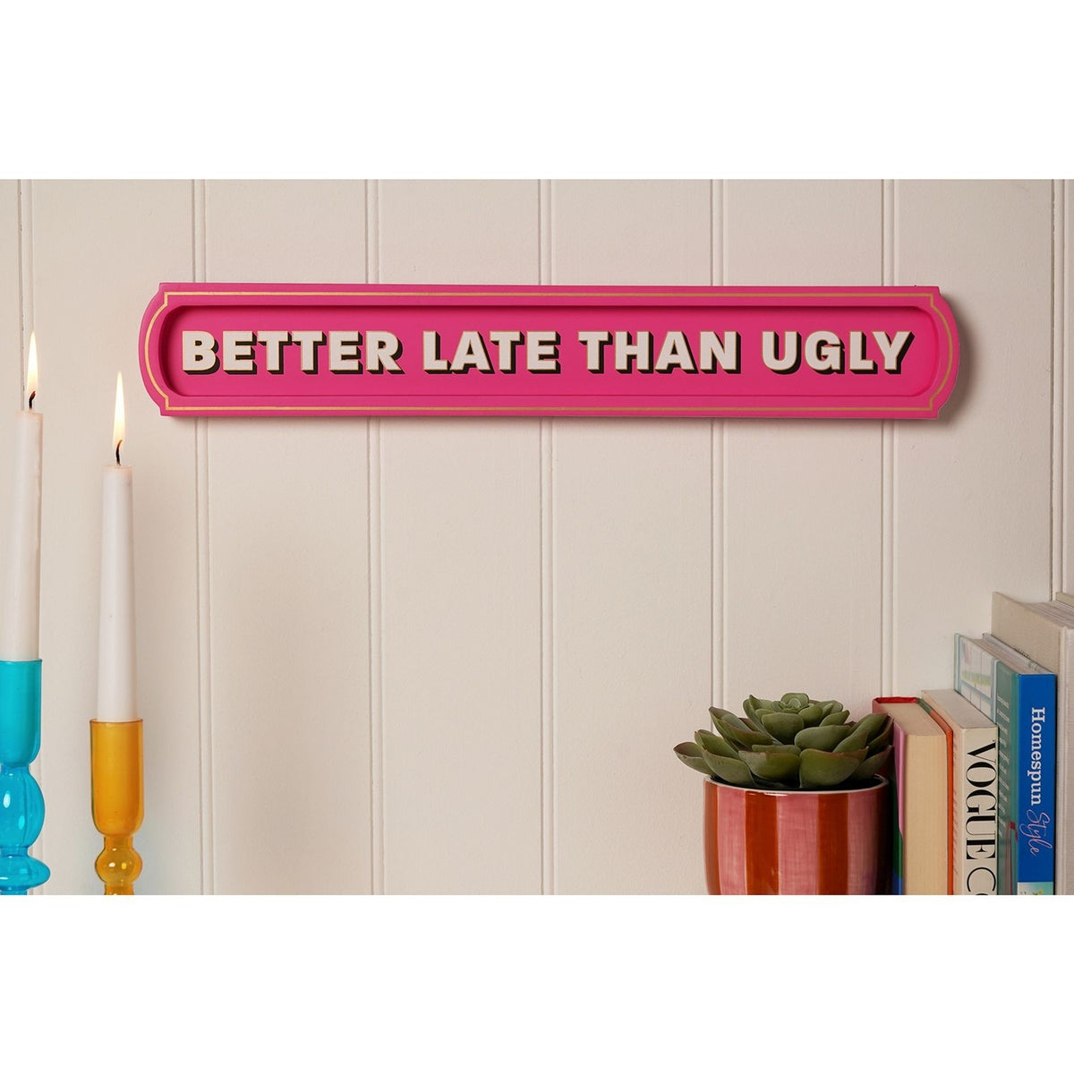 Pink wooden wall plaque reading "Better Late Than Ugly" in bold pale pink capitals with shadow detail, displayed on a panelled wall above a bookshelf and candles.
