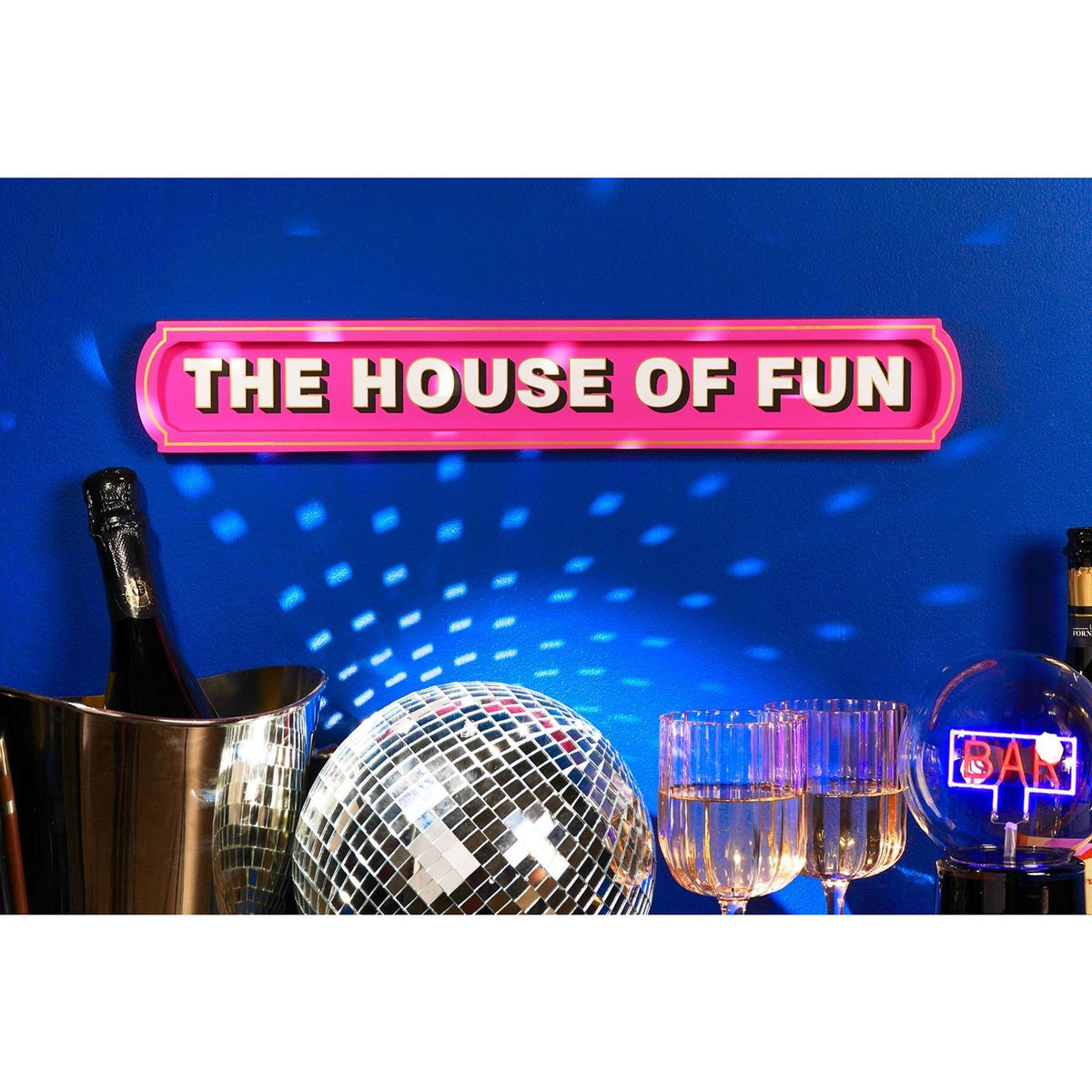 Pink wooden wall plaque reading "The House of Fun" in bold pale pink letters with black shadow and gold outline, mounted above a bar area with a disco ball and glassware.