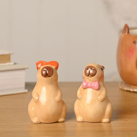 Capybara earthenware salt and pepper pot set, two tan ceramic figures with rosy cheeks, one wearing an orange bow and the other a pink bow tie, on a wooden surface.