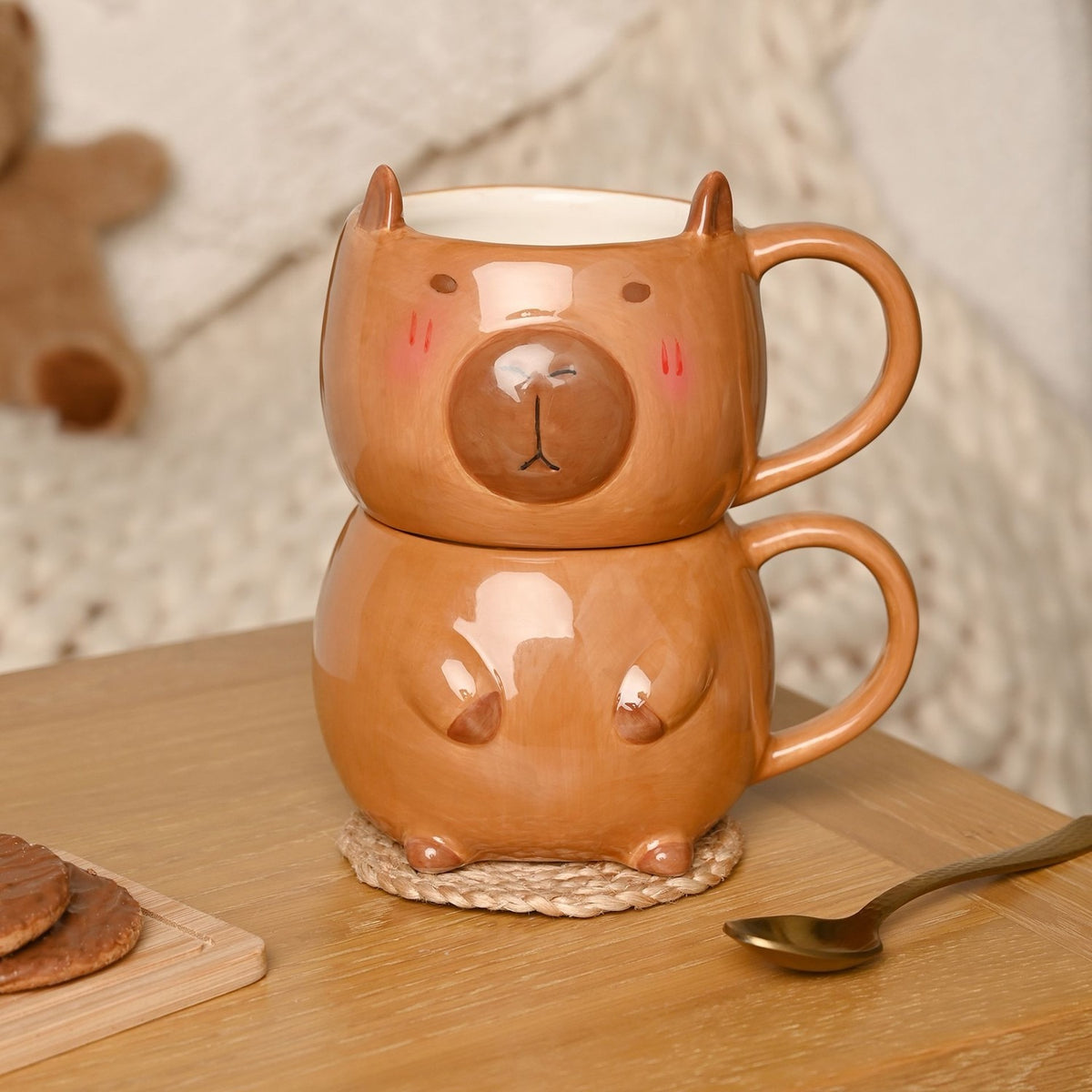 Glazed brown earthenware mug set shaped like a capybara head and body, stacked with loop handles, shown on a wooden table with biscuits and a spoon.