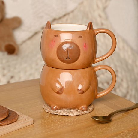 Glazed brown earthenware mug set shaped like a capybara head and body, stacked with loop handles, shown on a wooden table with biscuits and a spoon.