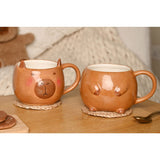 Two glazed earthenware capybara stacking mugs in brown, one with a face design and pointed ears, the other with paw details, shown on woven coasters on a wooden table.