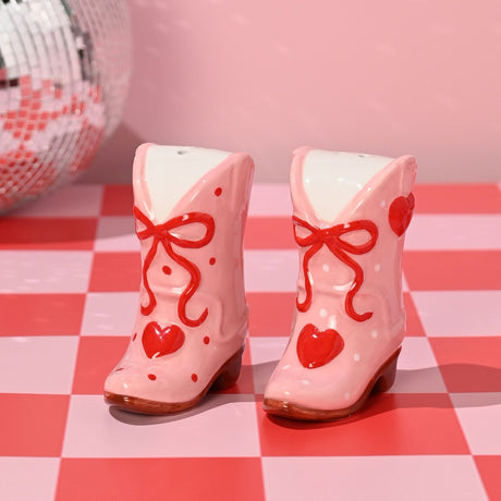 Pink ceramic cowboy boot salt and pepper set with red bows, hearts, and polka dot details, shown on a red and pink checkered surface.