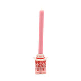 Ceramic taper candle holder shaped as pale pink cowboy boots with red heart and bow details beneath a white cowboy hat, holding a lit pink taper candle, on a plain white background.
