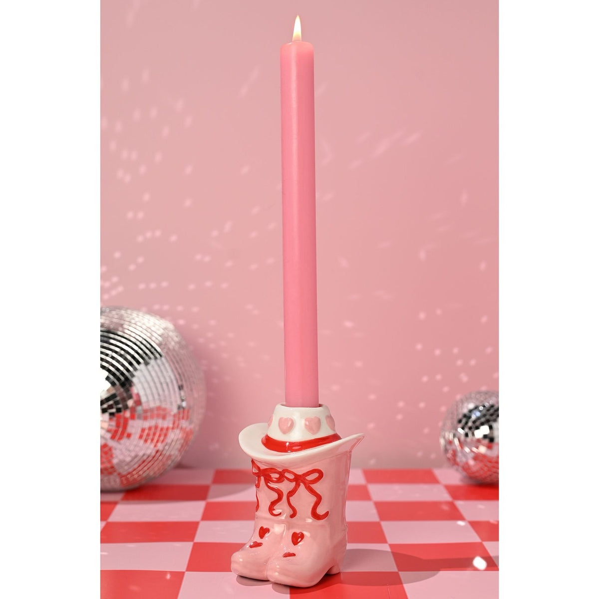 Ceramic Heart Cowboy Boot Taper Candle Holder in pink and white with red bows and hearts, holding a lit pink candle against a pink backdrop with disco balls.