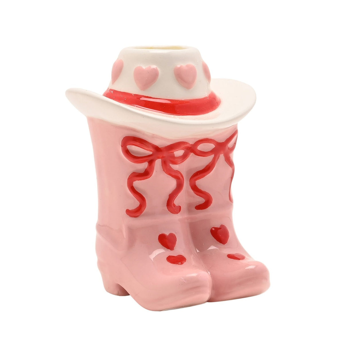 Ceramic taper candle holder shaped as pink cowboy boots with red bow laces and heart details, topped by a white cowboy hat with a red band and pink hearts, on a plain white background.