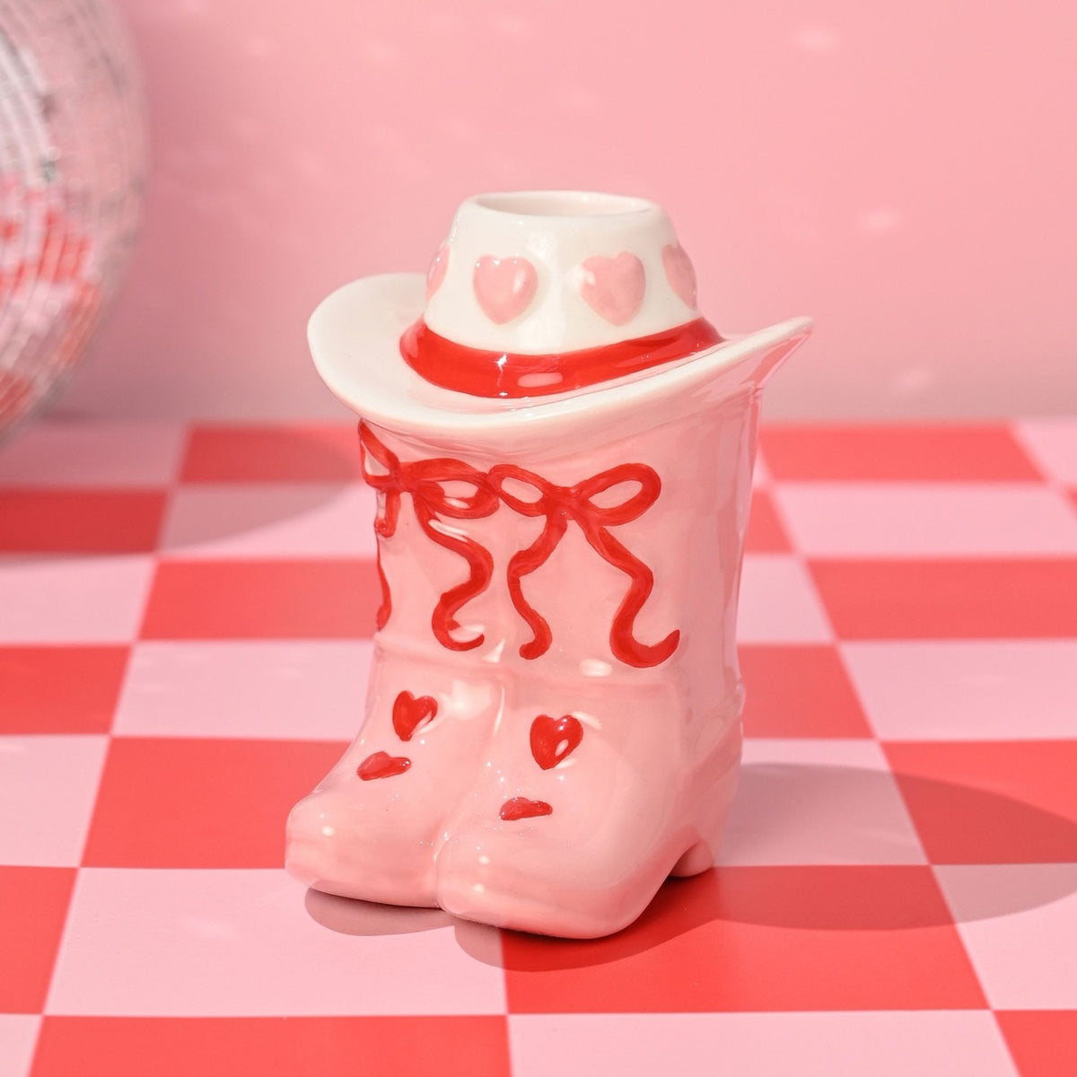 Ceramic taper candle holder shaped as pink cowboy boots with red heart and bow details, topped by a white hat with pink hearts and a red band, shown on a pink and red chequered surface.