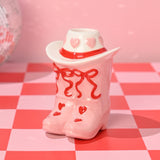 Ceramic taper candle holder shaped as pink cowboy boots with red heart and bow details, topped by a white hat with pink hearts and a red band, shown on a pink and red chequered surface.
