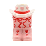 Ceramic cowboy boot taper candle holder in pink with a white hat, featuring red bows, pink and red hearts, and a glossy finish, on a plain white background.