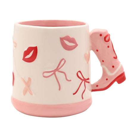 White earthenware mug with pink cowboy boot handle, featuring red and pink lips, bows, hearts and X motifs on a glossy white body with a pink base, shown on a plain background.