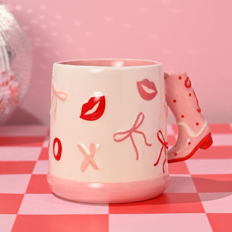 White ceramic Bow Earthenware Mug with pink cowboy boot handle, featuring raised lips, bows, and X and O motifs in pink and red, on a checkered surface.
