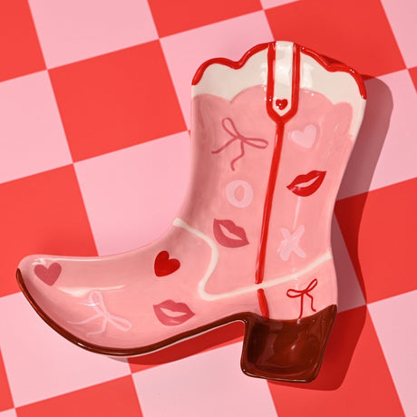 Cowgirl boot earthenware plate in pink with white top band, red seam and brown heel, decorated with hearts, lip prints and bows, on a pink and red checkered background.
