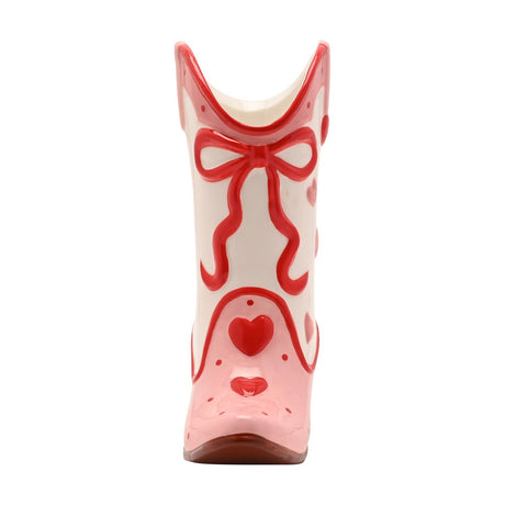 Ceramic cowgirl boot vase with cream shaft, pink cuff and toe, red piped outlines, a red bow at the top, and scattered red and pink hearts, on a plain white background.