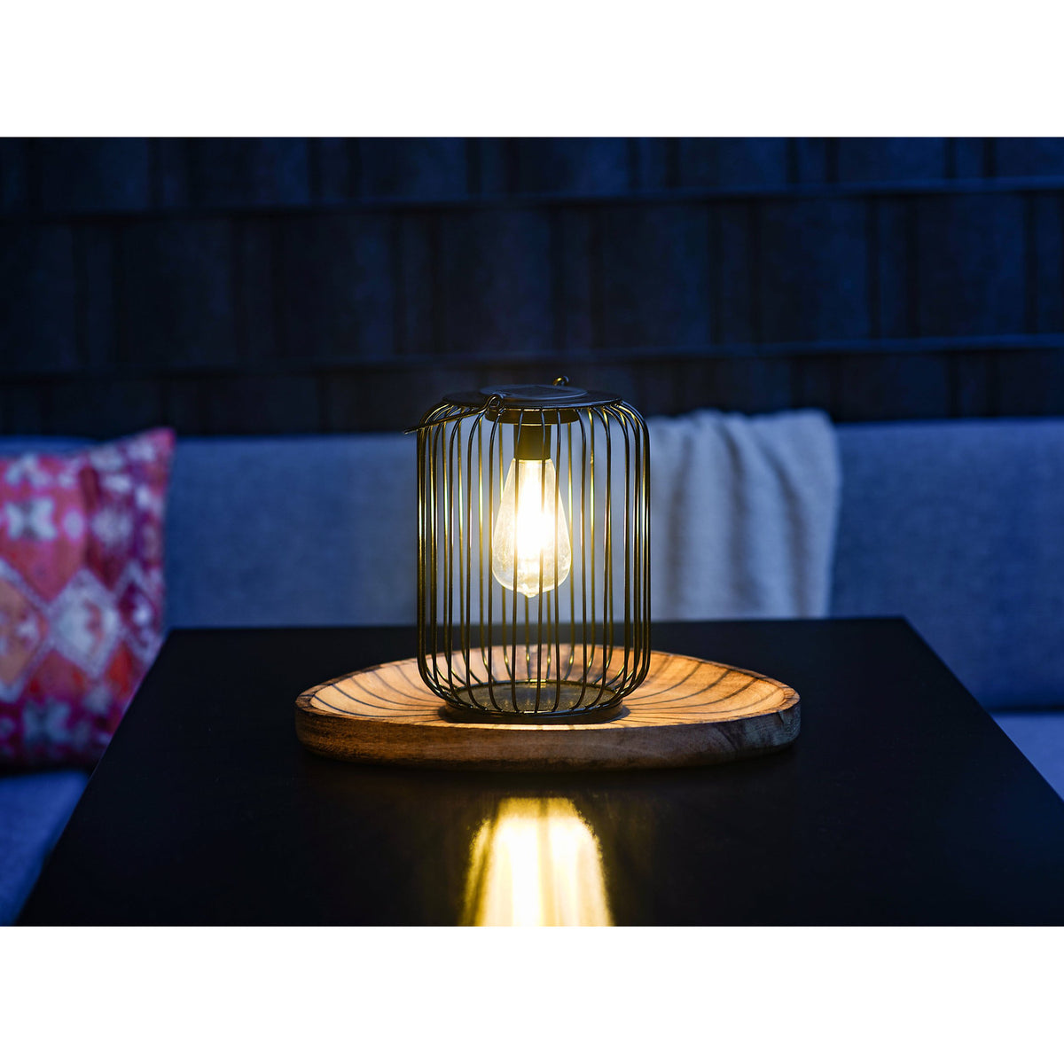 Solar Lantern 25cm in black steel wire with a rounded cage design and warm white LED filament-style bulb, lit on a wooden tray atop a dark table in a dim lounge setting.