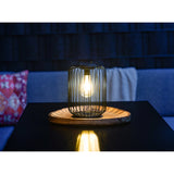 Solar Lantern 25cm in black steel wire with a rounded cage design and warm white LED filament-style bulb, lit on a wooden tray atop a dark table in a dim lounge setting.
