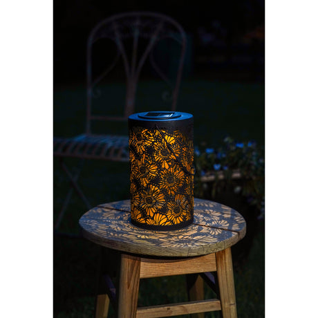 Solar LED table lamp with a cylindrical black cutout shade featuring a floral pattern, backed by a gold liner glowing warm white, shown lit on an outdoor wooden stool at night.