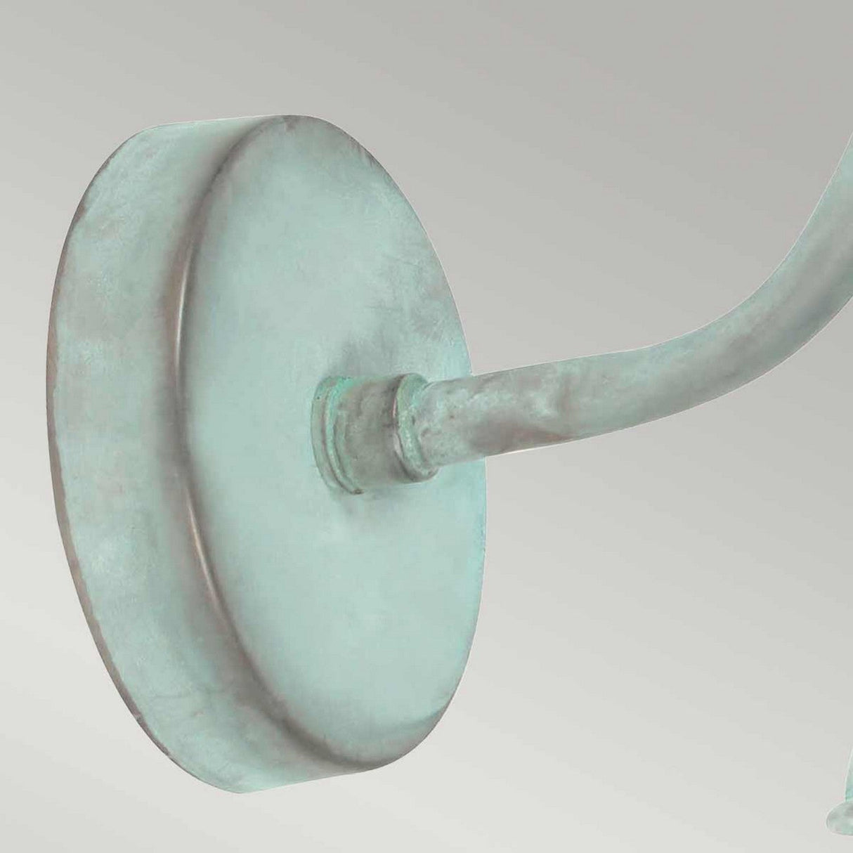A close-up view of the Somerton 1 Light Outdoor Wall Light highlights its greenish verdigris metal mount, featuring a round base and curved arm that suggests it might be crafted from bronze or copper, adding an air of elegance and age.