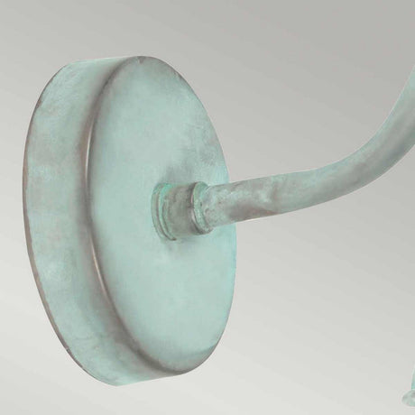 A close-up view of the Somerton 1 Light Outdoor Wall Light highlights its greenish verdigris metal mount, featuring a round base and curved arm that suggests it might be crafted from bronze or copper, adding an air of elegance and age.
