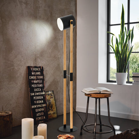Sonanne wooden floor lamp with pale wood uprights, black drum shade, and round black base, shown beside a stool and plant in a living room.
