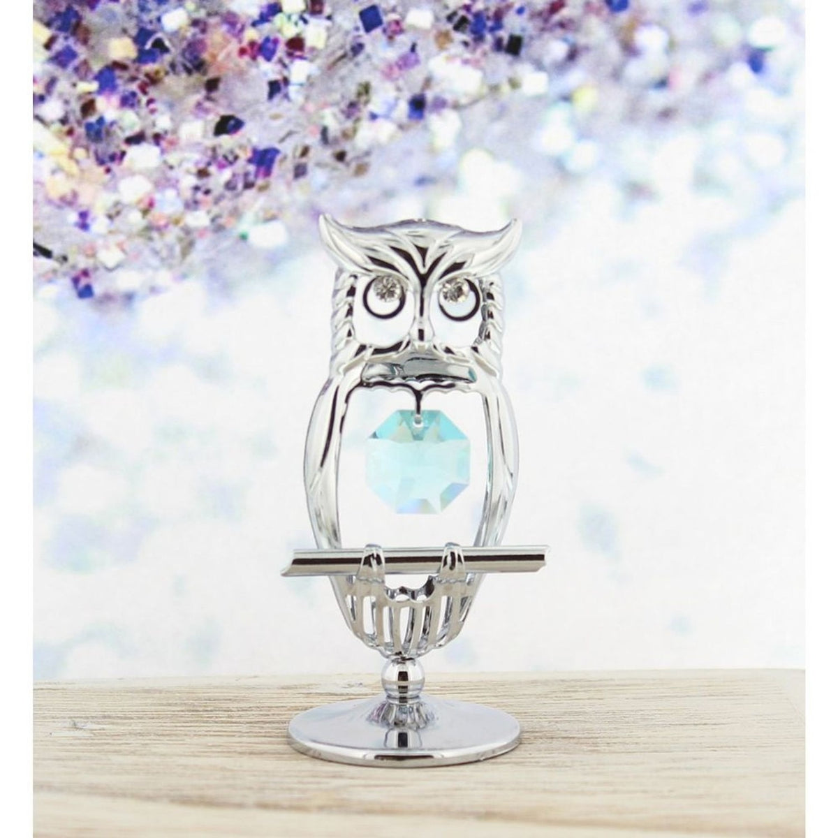 Chrome plated perched owl figurine with open silver outline body, tufted ears, crystal stone eyes, and a faceted light blue crystal held at its centre, on a round pedestal base.